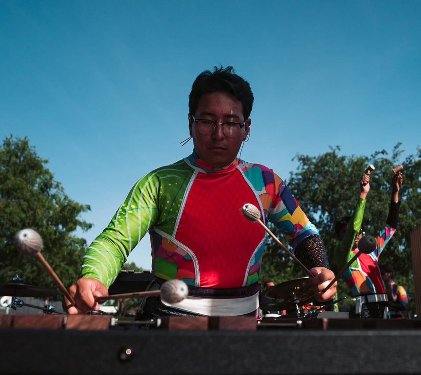 Person playing a large outdoor percussion instrument with colorful clothing against a blue sky.